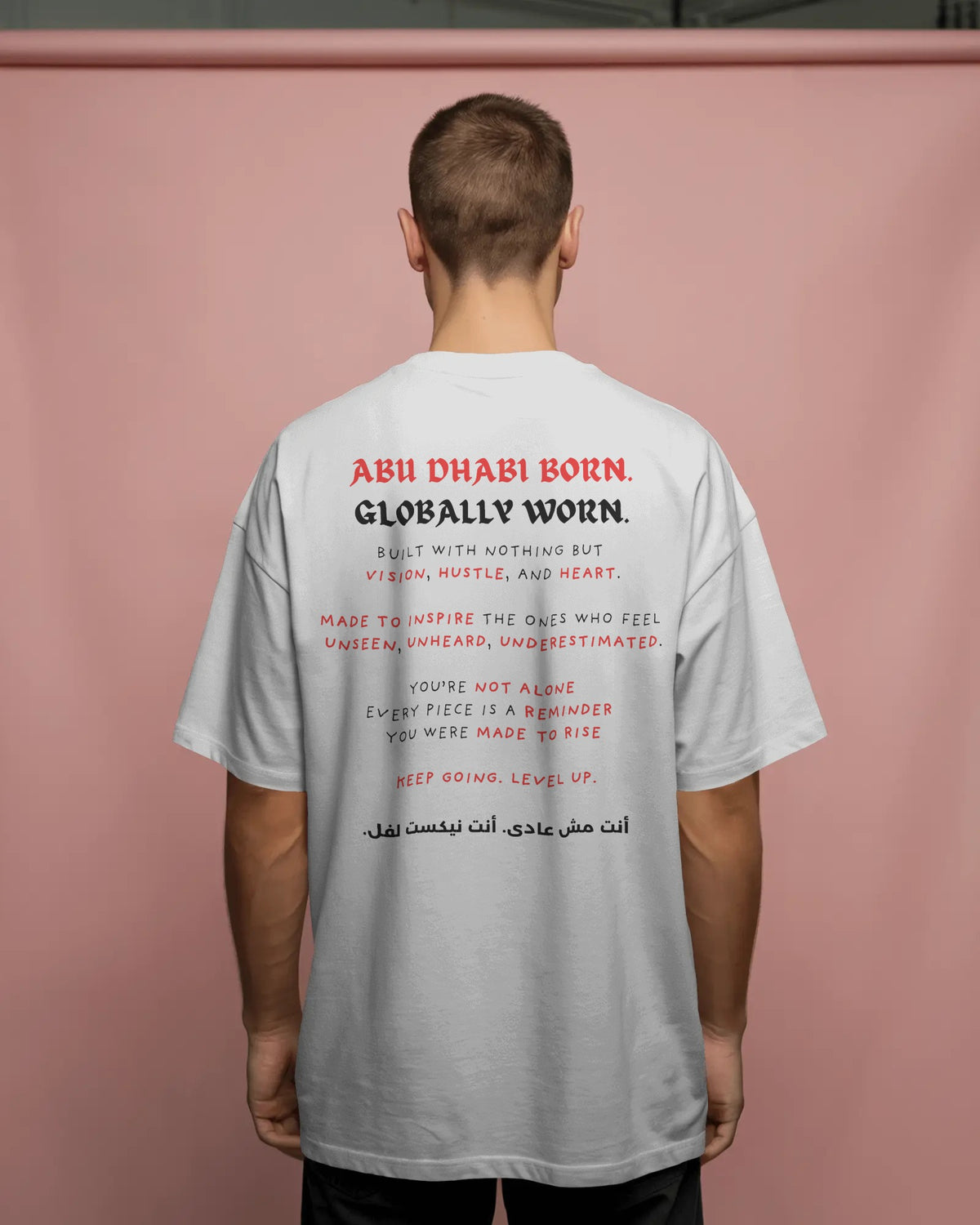 Back view of NXT LVL "Abu Dhabi Born. Globally Worn" limited drop tee on male model against pastel pink studio background
